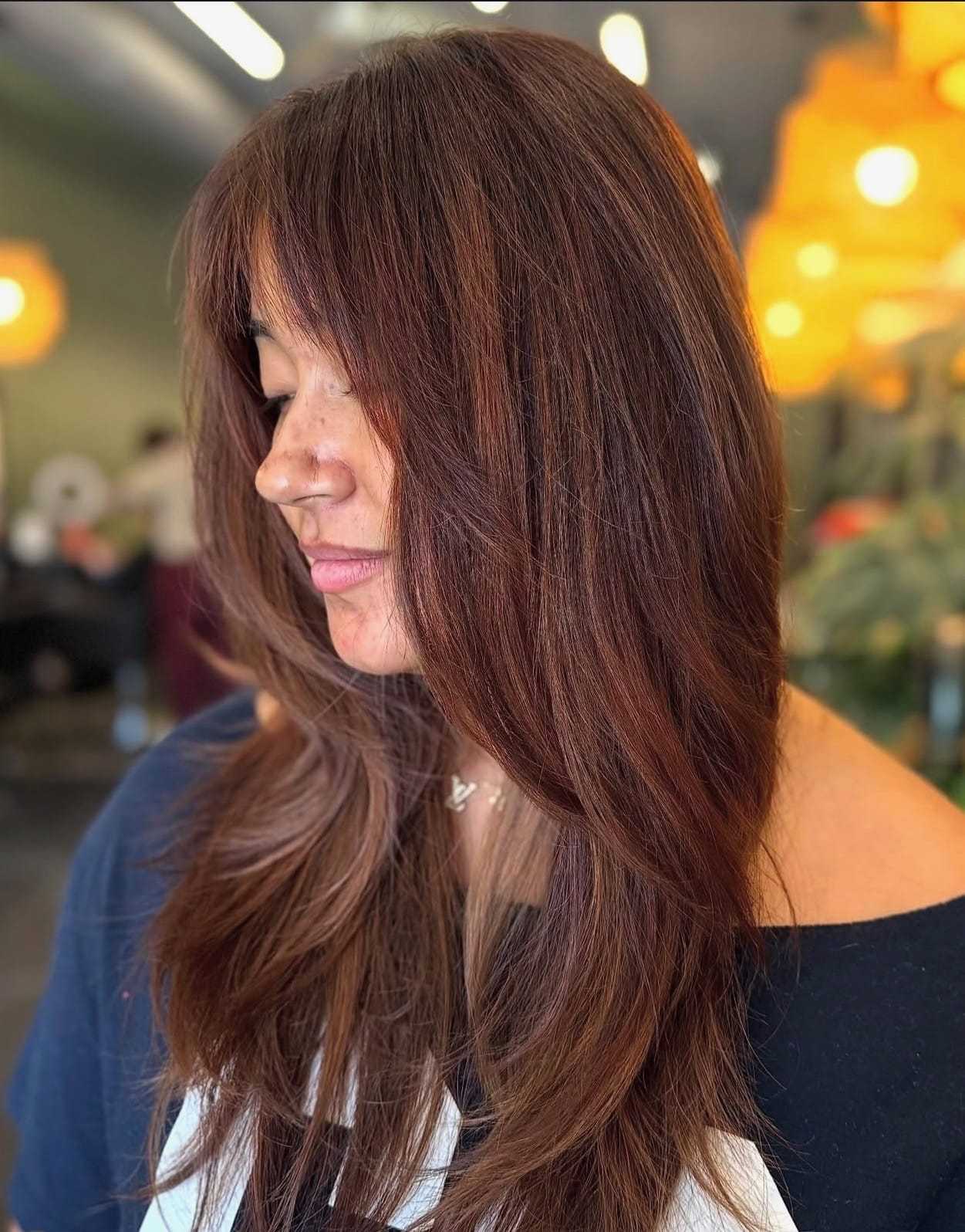 Woman with layered brown hair and bangs, wearing a dark shirt, in softly lit interior.