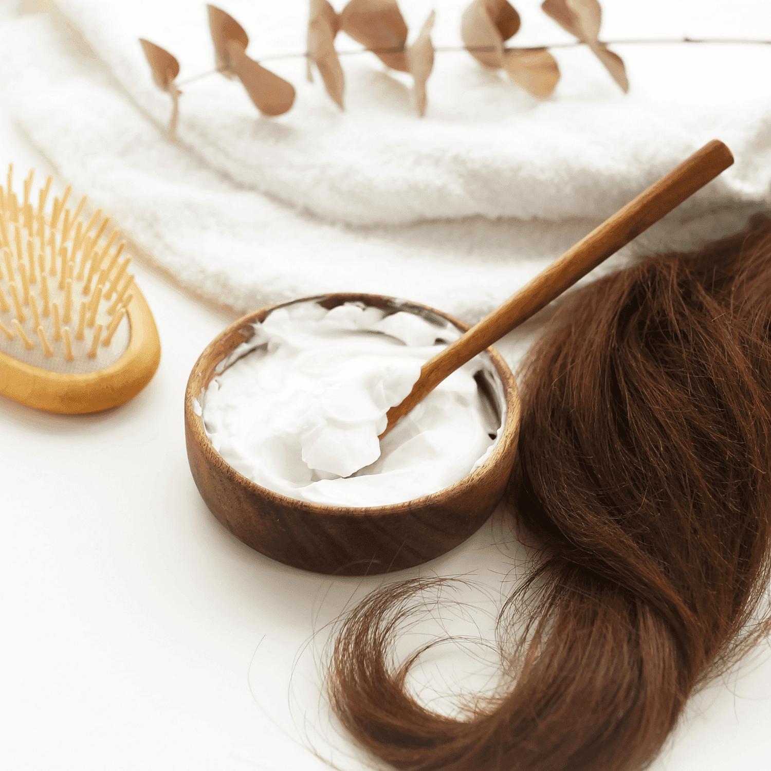 Hair mask in wooden bowl with brush, brown hair, and towel on white background.